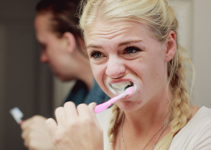 Young woman brushing teeth intensely in bathroom with another person blurred in background, showcasing unusual wellness habits