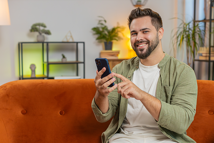 Man happy holding phone on orange couch, smiling confidently after ex-wife caught cheating in modern living room.
