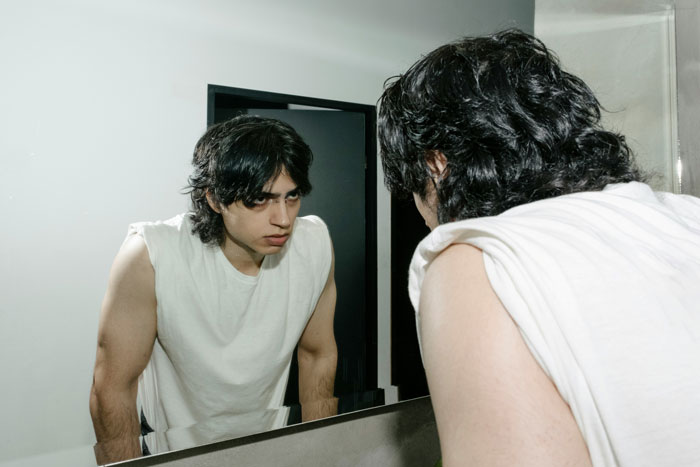 Young man in white sleeveless shirt looking intently at himself in the bathroom mirror, reflecting on life challenges.