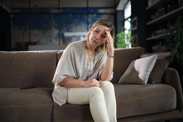 Worried woman sitting on couch looking thoughtful, reflecting on life challenges and seeking comfort with wine.