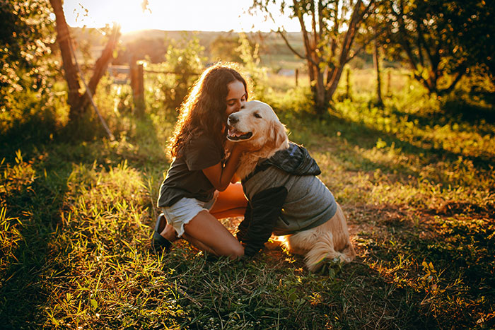 Woman with service dog outdoors at sunset, sharing a tender moment during a peaceful autumn setting.