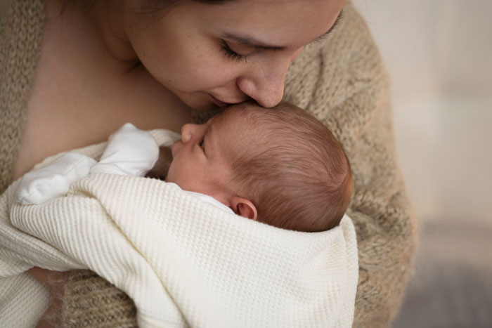 Indian grandmother holding newborn baby wrapped in white blanket, looking closely at the child in a cozy setting.