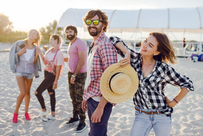 Man spending holiday closely with wife&rsquo;s friend on beach, while others stand in background in casual summer attire.