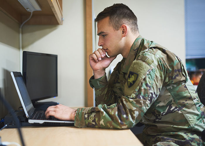 Man in military uniform looking at laptop screen, appearing thoughtful while working indoors.