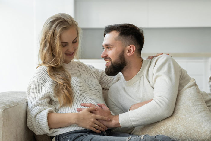 Couple sitting on couch, woman touching her belly while man smiles, relating to pregnancy obsession and health concerns.