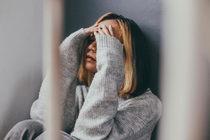 Woman in gray sweater sitting against wall covering face, representing obsession and life-threatening condition concerns.