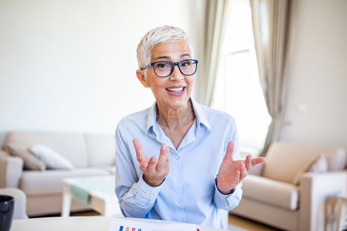 Senior woman with glasses discussing husband retirement and children finances at home, smiling and explaining with hand gestures.