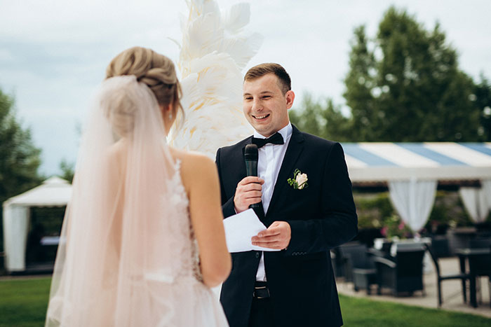 Groom in tuxedo holding a microphone and reading vows to bride during emotional outdoor wedding ceremony. Groom in tuxedo holding a microphone and reading vows to bride during emotional outdoor wedding ceremony.