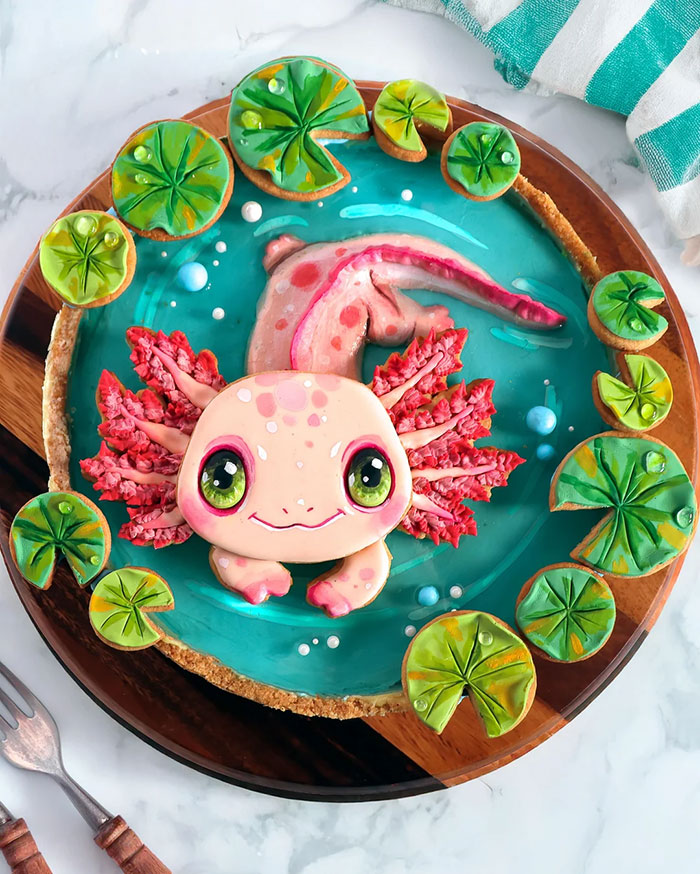 Axolotl-themed baked goods with colorful icing and detailed lily pad decorations on a round wooden board.