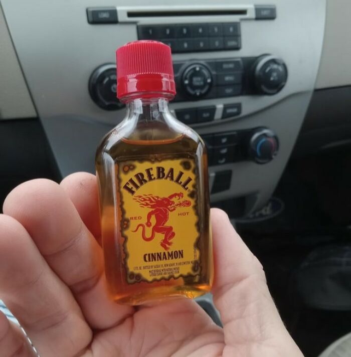 Hand holding a small Fireball Cinnamon whiskey bottle inside a car, illustrating Thanksgiving arguments about mashed potatoes and drinks.