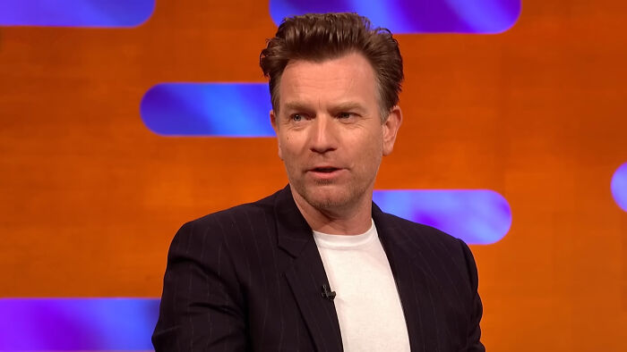 James Franco in a dark blazer and white shirt speaking during a TV interview with an orange and blue background.