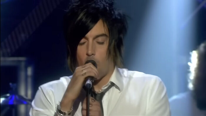 Male singer with dark hair and white shirt holding a microphone under dramatic stage lighting, James Franco smells theme.