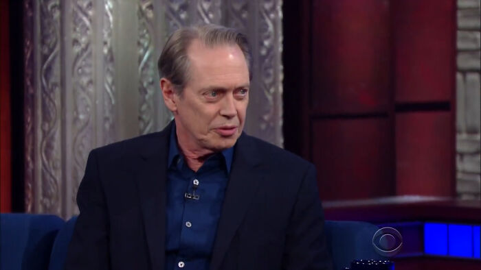 Steve Buscemi in a talk show setting, wearing a dark suit and blue shirt, engaged in conversation on set.