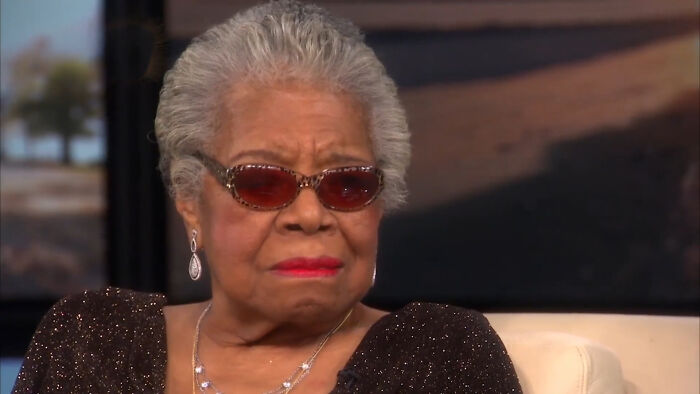 Elderly woman wearing red sunglasses and a sparkly black dress, seated indoors with a neutral expression.