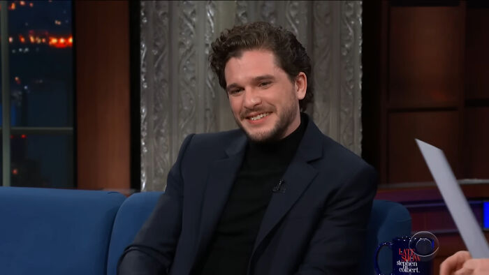 Actor James Franco smiling on a talk show set, wearing a black turtleneck and dark blazer during an interview.