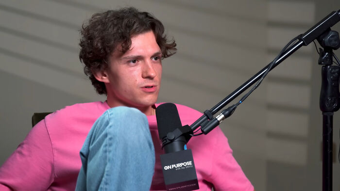 Tom Holland in a podcast studio, wearing pink shirt and jeans, speaking into a microphone about James Franco smells.