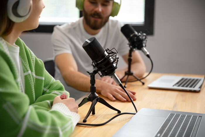 Two people wearing headphones recording a podcast with microphones and laptops on a wooden table discussing James Franco smells.