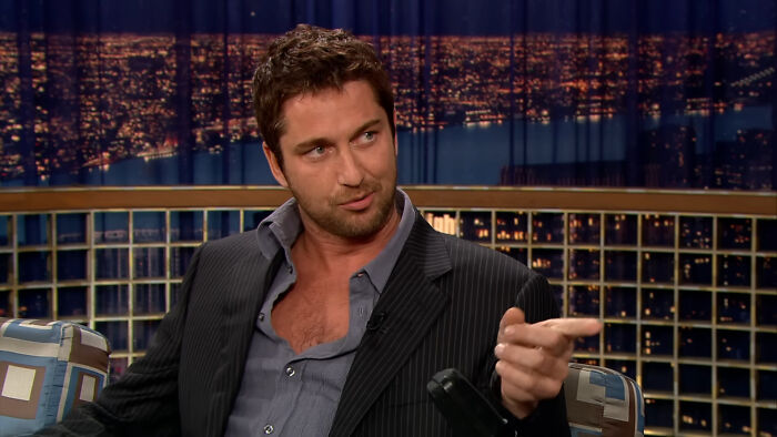 James Franco in a dark suit sitting on a talk show set with city lights in the background, gesturing with his hand.