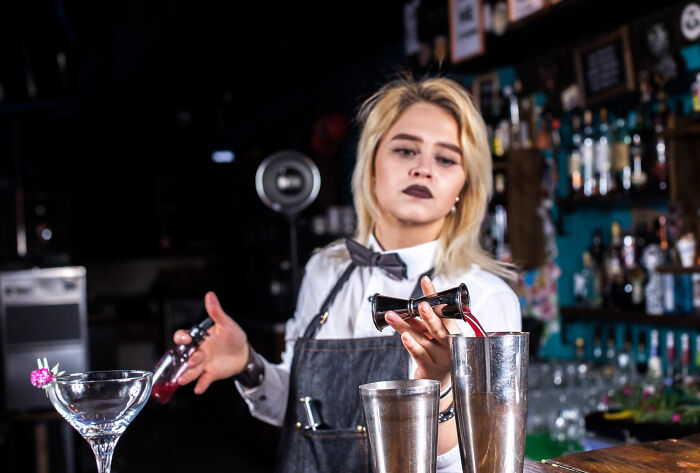 Bartender pouring a red drink from a jigger into a shaker in a dimly lit bar with shelves of bottles behind her.