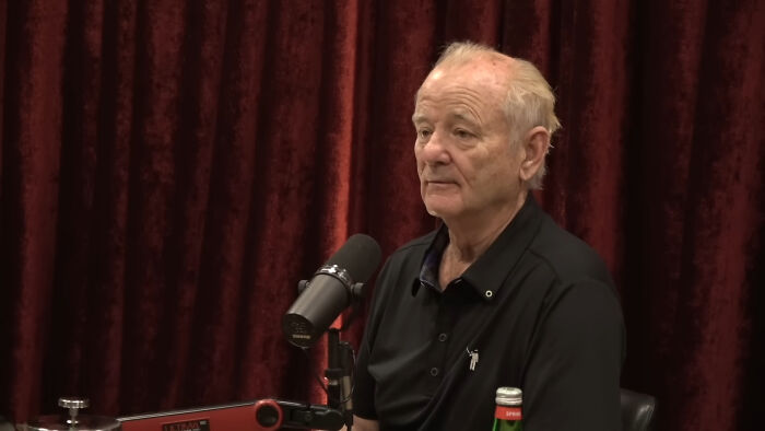 Older man in a black shirt sitting by a microphone in a podcast studio with a red curtain background, discussing James Franco smells.