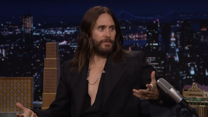 James Franco speaking on a late-night talk show set, wearing a black blazer with a necklace, gesturing with his hands.