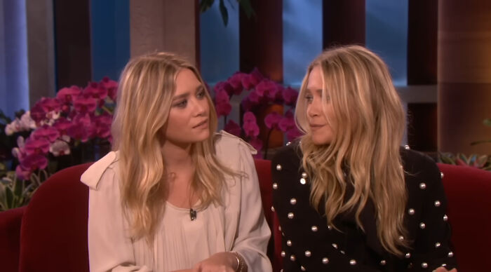 Two women with long blonde hair sitting on a red couch, engaged in conversation, with purple flowers in the background.