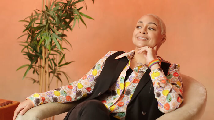 Person with short blonde hair wearing a colorful patterned shirt and black vest seated in a chair, smiling confidently.