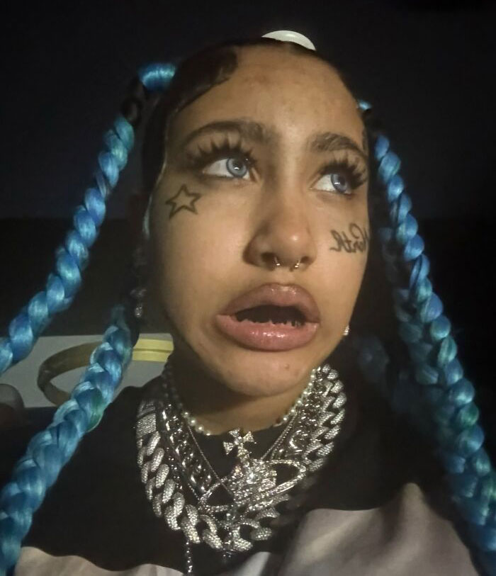 Person with blue braided hair and facial tattoos wearing layered chains, amid celebrity parenting debate and backlash over North West comparison.