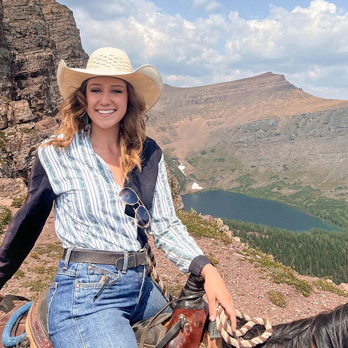 Young woman wearing cowboy hat and striped shirt sitting on horseback with mountain and lake landscape behind, karma train keyword included.