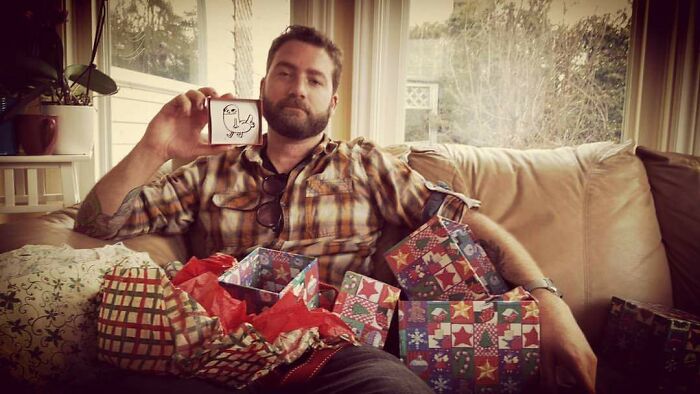 Man sitting on couch surrounded by holiday gift boxes holding a playful drawing showing men never grow up.
