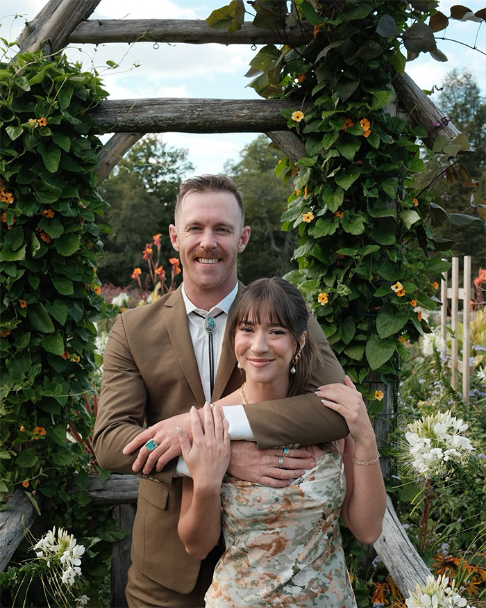 Man and woman posing outdoors surrounded by flowers and greenery, representing Jiu-Jitsu trainer accident context.