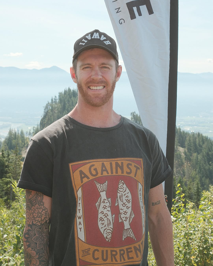 Jiu-Jitsu trainer outdoors wearing a black Against The Current t-shirt and Hawaii cap with mountain landscape background.