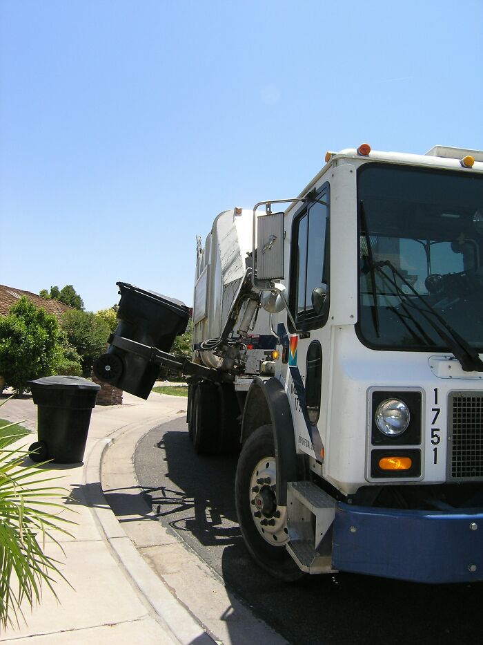 Garbage truck lifting a black trash bin on a suburban street during clear weather in garbage men horror job scenario.