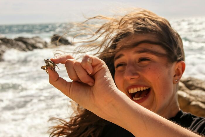 Young woman holding a small crab by the ocean, illustrating people explain the etymology of common words concept.