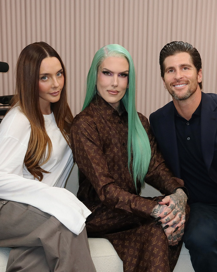 Jeffree Star seated between two people, wearing a brown patterned outfit with green hair and makeup, posing indoors.