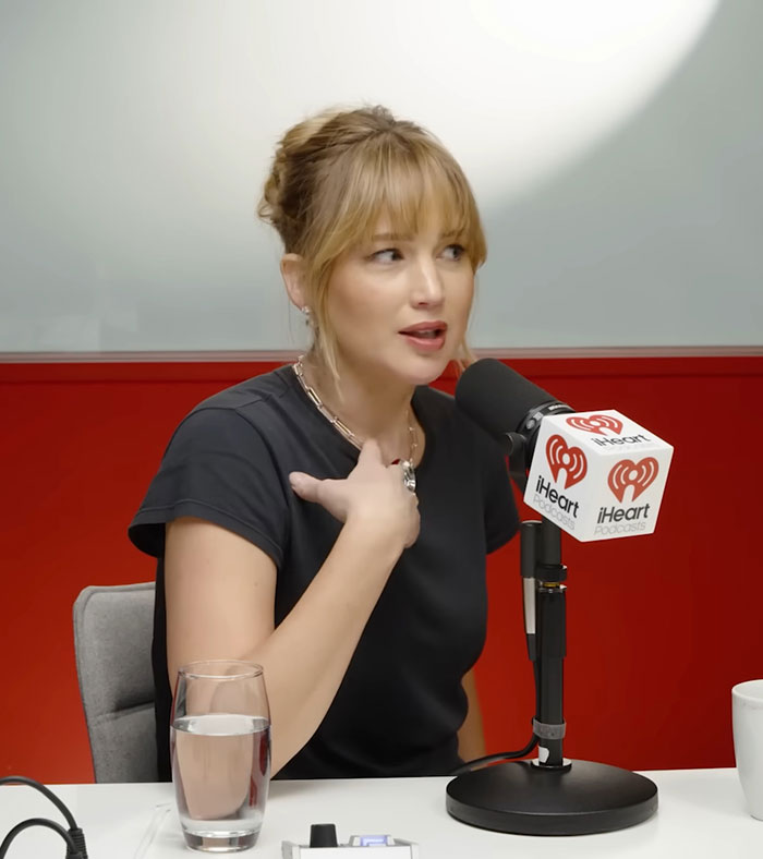 Jennifer Lawrence speaking into an iHeart podcast microphone during an interview with a glass of water on the table.