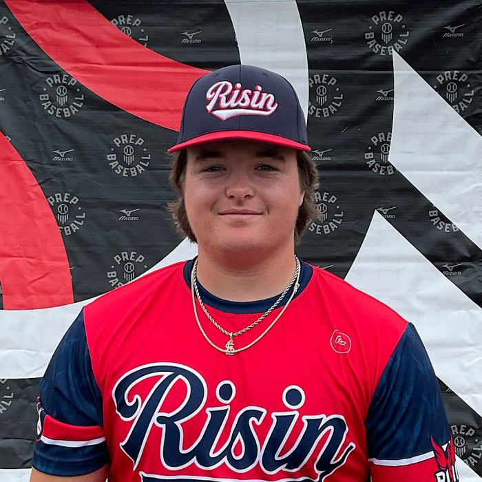 Teen wearing Risin baseball uniform and cap standing in front of a banner at a baseball event in Oklahoma protests.