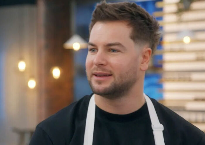 Chris Hughes wearing a black shirt and white apron, appearing thoughtful during a reality TV show confession.