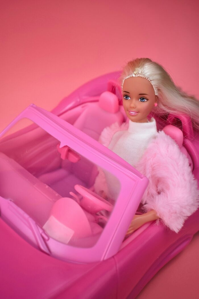 Blonde doll dressed in pink driving a pink toy car, representing a Karen character in a playful setting.