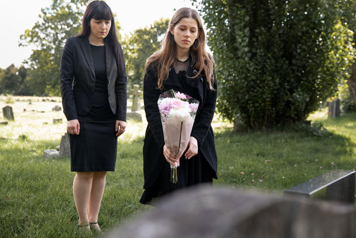 Two women dressed in black mourning at a graveyard, one holding flowers, reflecting a widowed friend and family conflict. Two women dressed in black mourning at a graveyard, one holding flowers, reflecting a widowed friend and family conflict.