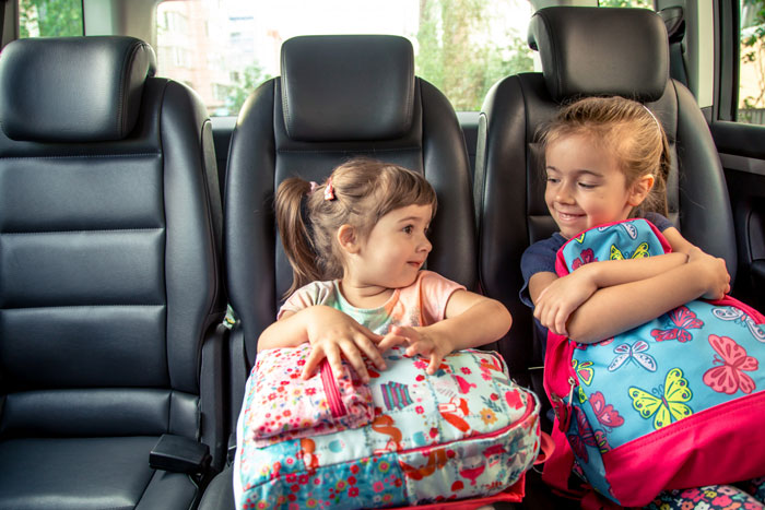 Two young girls sitting in a car backseat holding colorful backpacks showing family and mom prioritizes widowed friend concept. Two young girls sitting in a car backseat holding colorful backpacks showing family and mom prioritizes widowed friend concept.