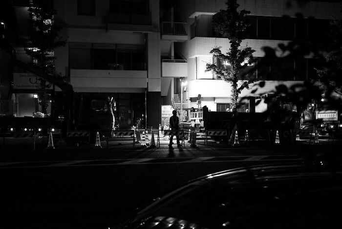 Dark urban scene captured by surveillance cameras showing shadowy figures near construction cones at night.