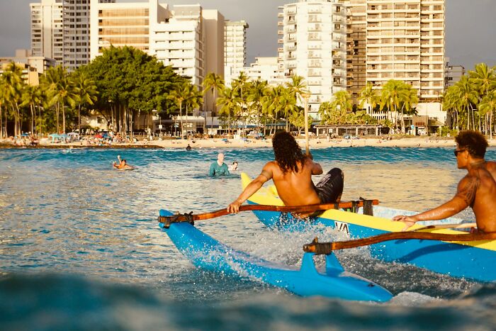 Two men paddling a blue outrigger canoe in Miami waters with city buildings and palm trees in the background.