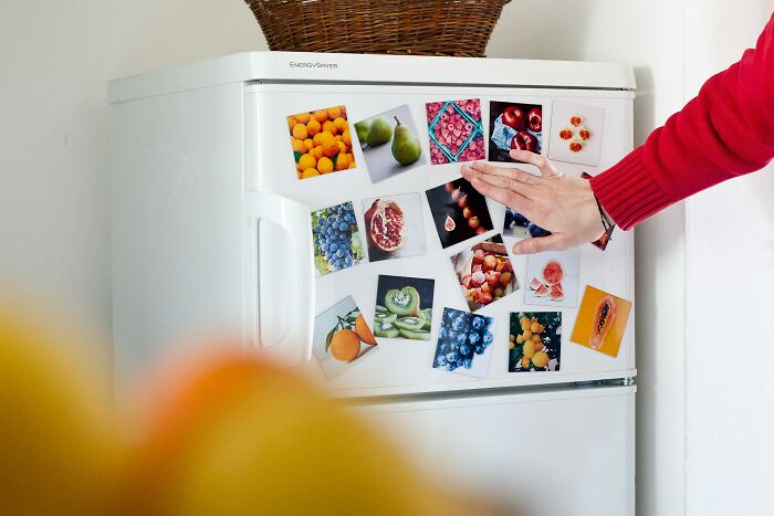 Hand placing photo magnets on fridge covered with colorful fruit images, capturing funny and serious parenting moments.