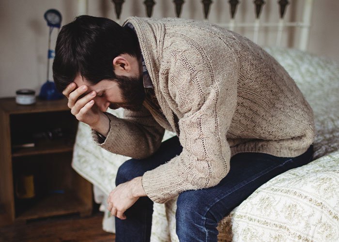 Man in beige sweater sitting on bed, holding head in hand, reflecting on things learned embarrassingly late in life.