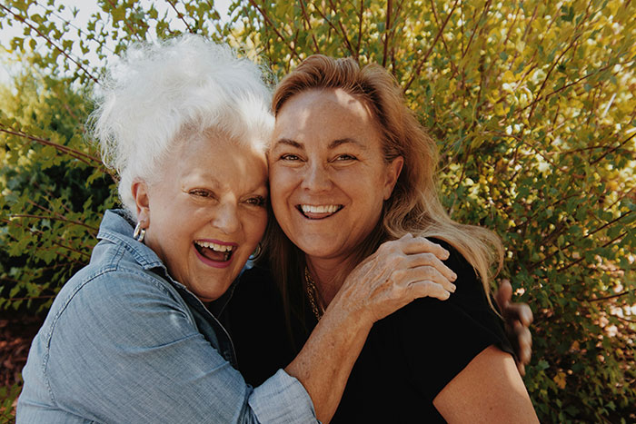 Two women smiling and hugging outdoors with sunlight and green foliage, highlighting woman and service dog themes.