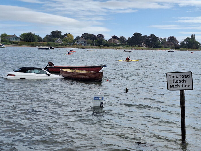 Partially submerged car near boats and flooded road sign, illustrating zero common sense in a flooded area.