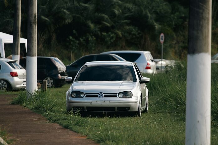 White Volkswagen car parked on grass near a sidewalk surrounded by other vehicles in an outdoor area.