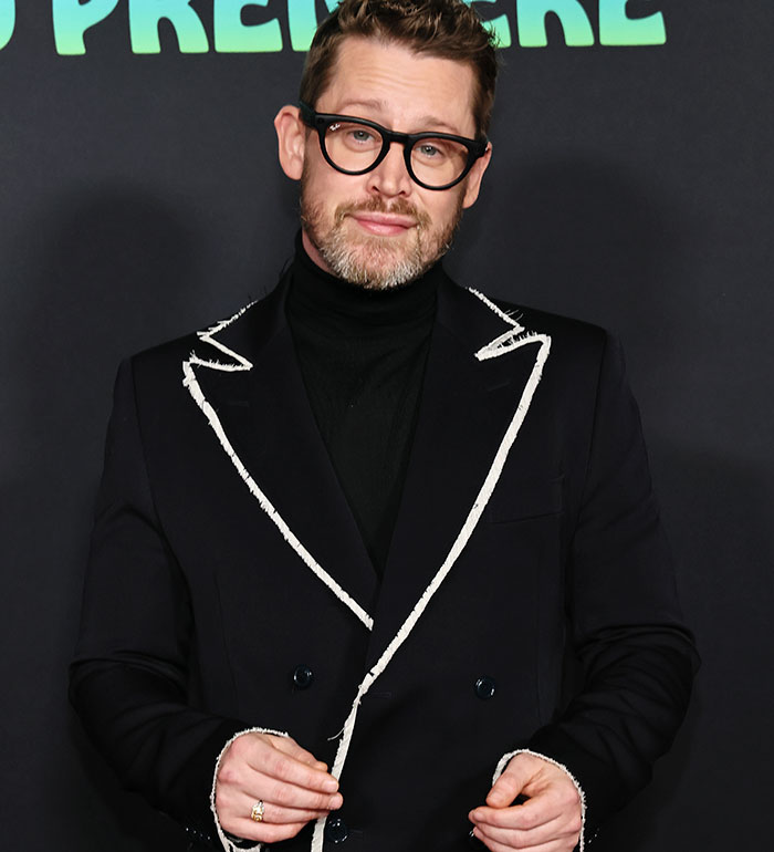 Macaulay Culkin wearing glasses and a black suit with white trim at a premiere event, looking confident and relaxed