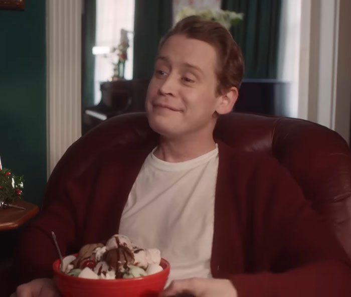 Macaulay Culkin sitting in a chair smiling while holding a bowl of ice cream, casually dressed in a white shirt and cardigan.